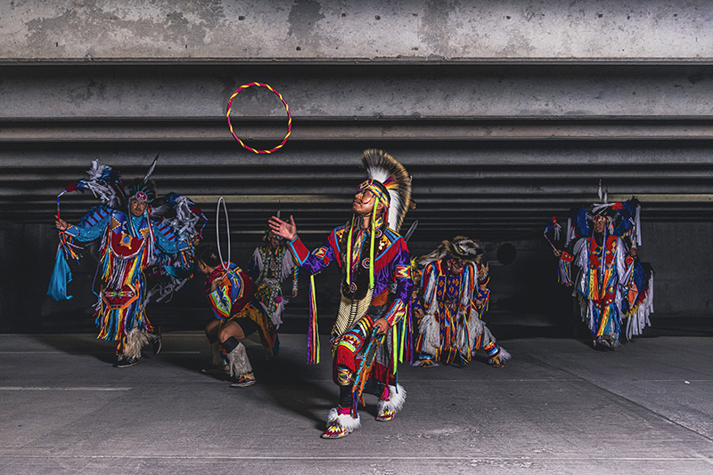 Six members of dance troupe Indigenous Enterprise perform in colorful regalia, against a concrete background.