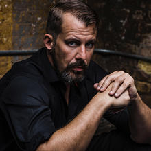 Baritone Kyle Albertson, with brown hair, mustache, and beard, sits with his hands crossed, wearing a black, short-sleeved shirt.