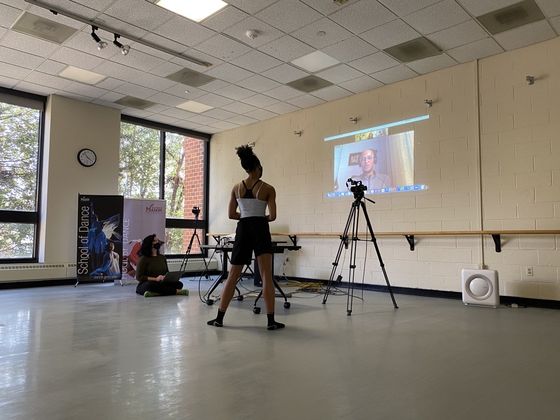 Olivia McCall, a young Black woman, stands with her back to the camera, looking at the projection of a Zoom call with Rafael Palacios, the founder of Sankofa Danzafro. Setarra Kennedy, faculty mentor, sits on the floor to the left of McCall..