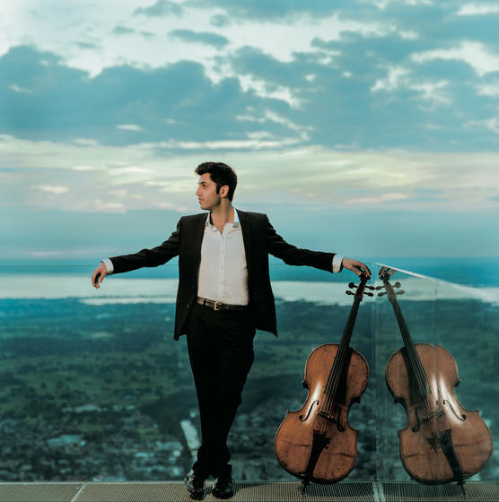 Kian Soltani, dressed in a suit, stands in front of blue sky with a cello to his left.