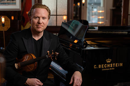 Sitting at a black grand piano, red-haired violinist Daniel Hope holds his violin and smiles.